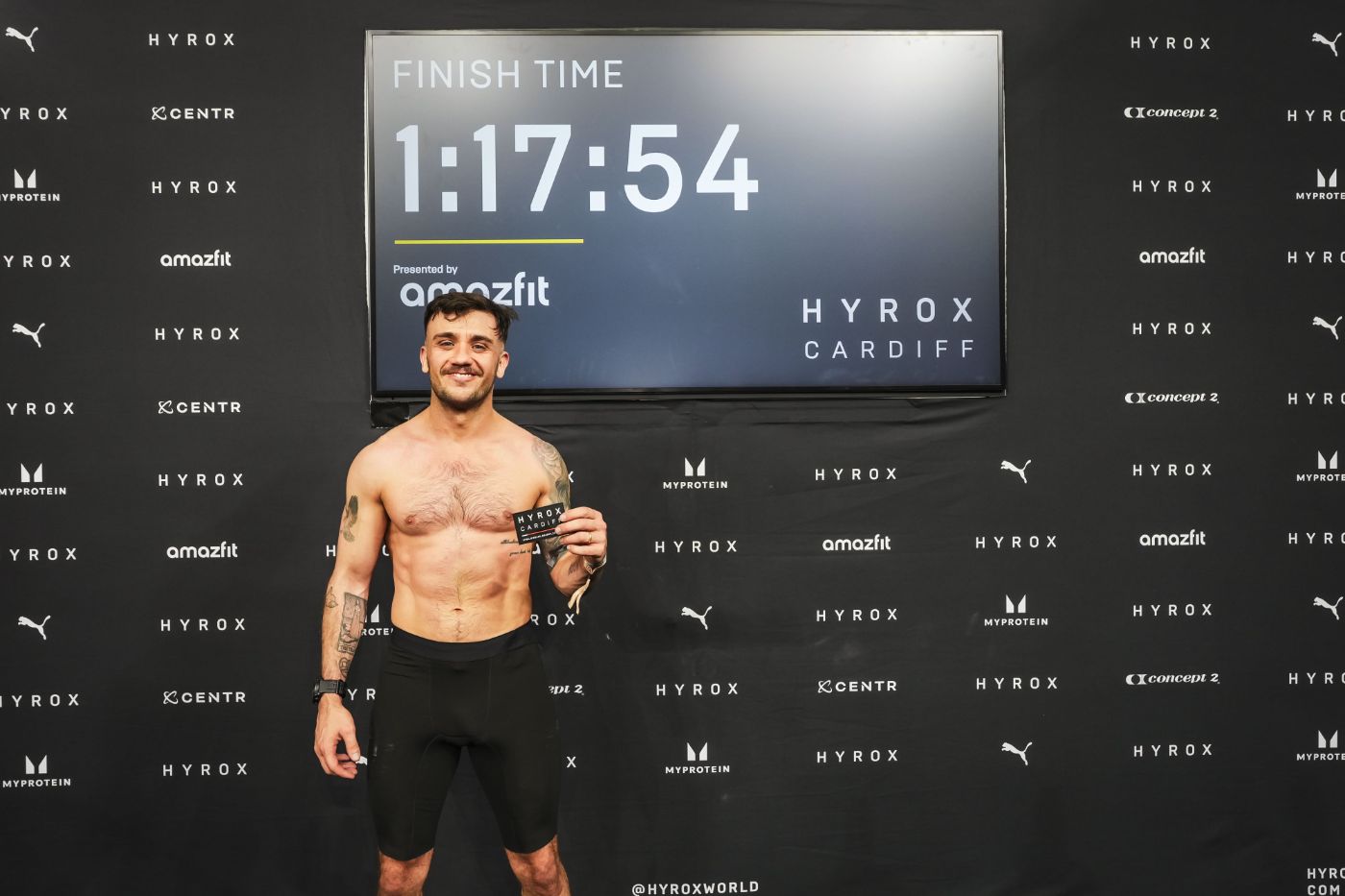 Reece holding his finisher card in front of the HYROX Cardiff screen showing 1:17:54